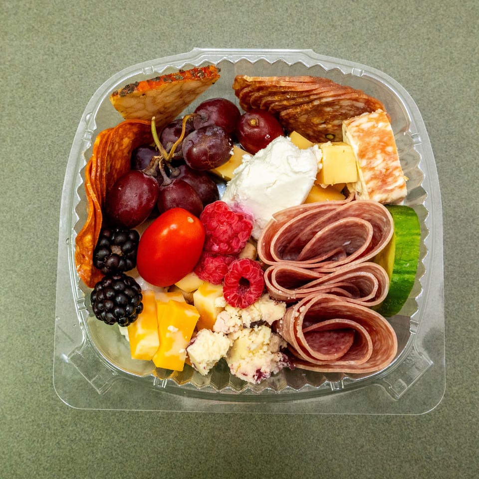 Charcuterie platter with folded salami, cheeses, crackers, grapes, berries, tomatoes, cucumber, and raspberries in a clear container.