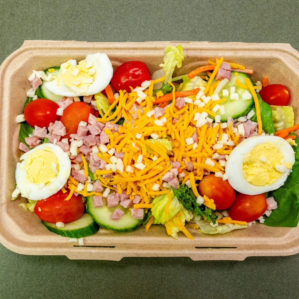 Chef salad with lettuce, ham, shredded cheddar, hard-boiled eggs, tomatoes, cucumbers, carrots, and onions in a takeout container.